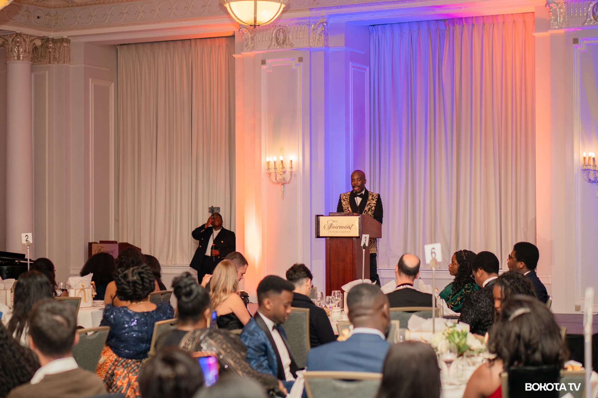 A speaker in a gold-embroidered jacket addresses guests from the Fairmont Château Laurier podium during MBOMBO Gala Night