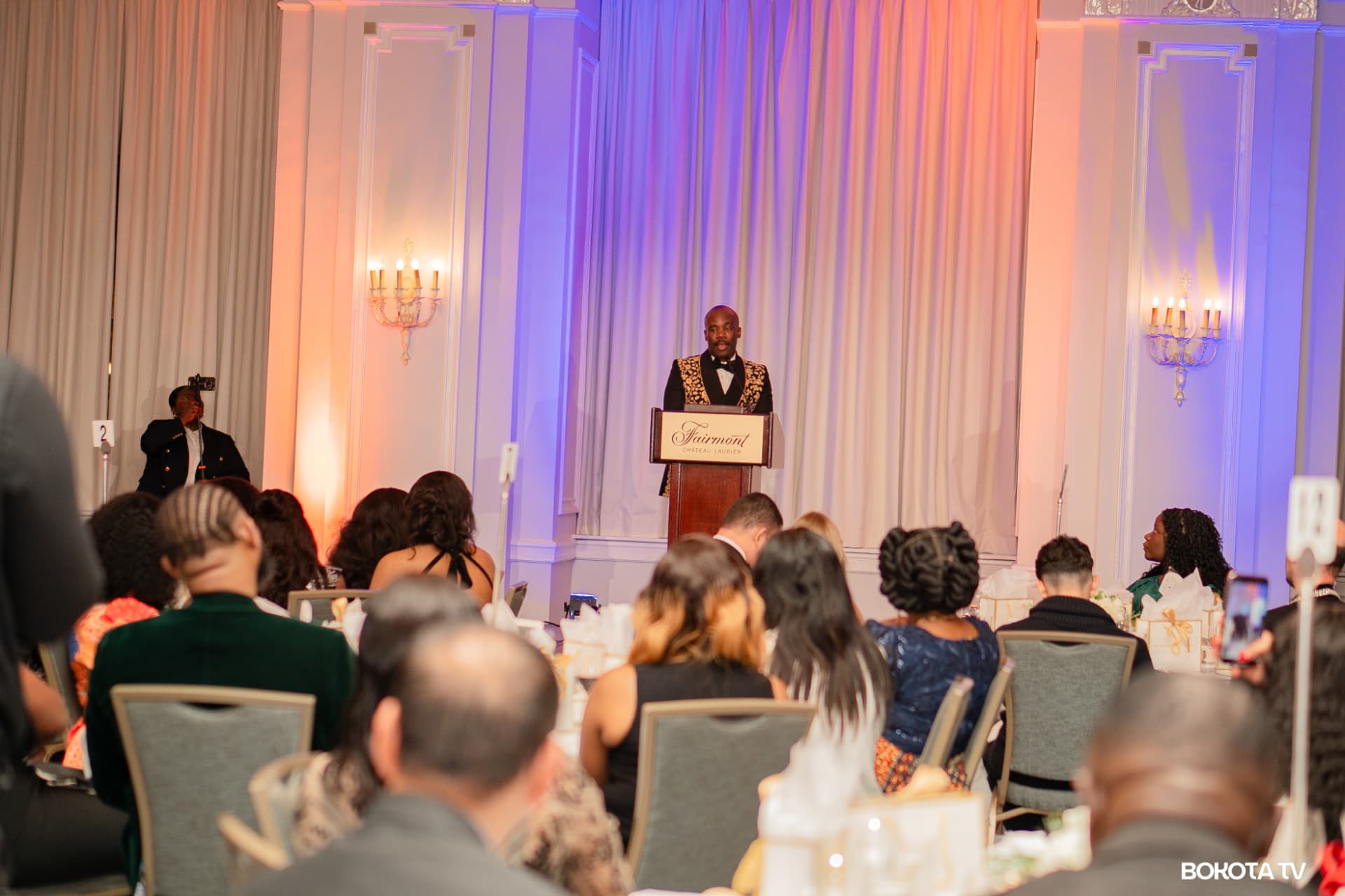 The MC at the Fairmont Château Laurier podium in the Château Laurier ballroom, lit in warm amber and violet